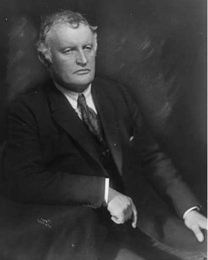 Edvard Munch by Anders Beer Wilse (1921) - Black and white portrait of Edvard Munch in a suit in 1921, seated against a dark backdrop. His expression is serious, with a focused and introspective look.