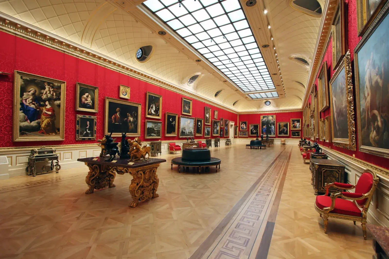 The Great Gallery at the Wallace Collection: a long room with red damask walls hung with gilt-framed Old Master paintings, a glazed skylight running the length of the ceiling, and inlaid furniture on a parquet floor